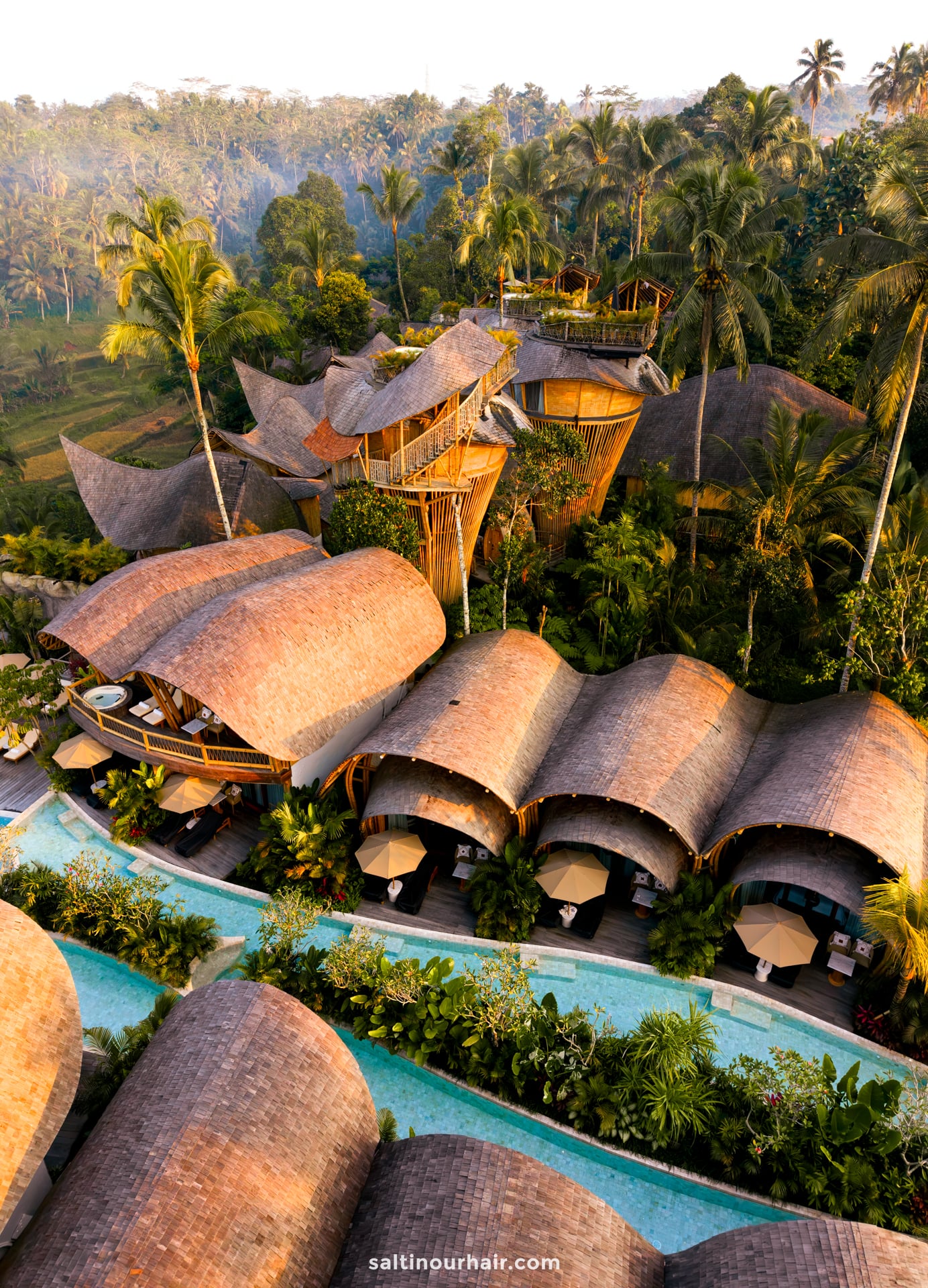 Aerial view of a tropical bambootel bamboo hotel bali with thatched-roof buildings, surrounded by lush greenery and palm trees, featuring a swimming pool and shaded lounge areas.