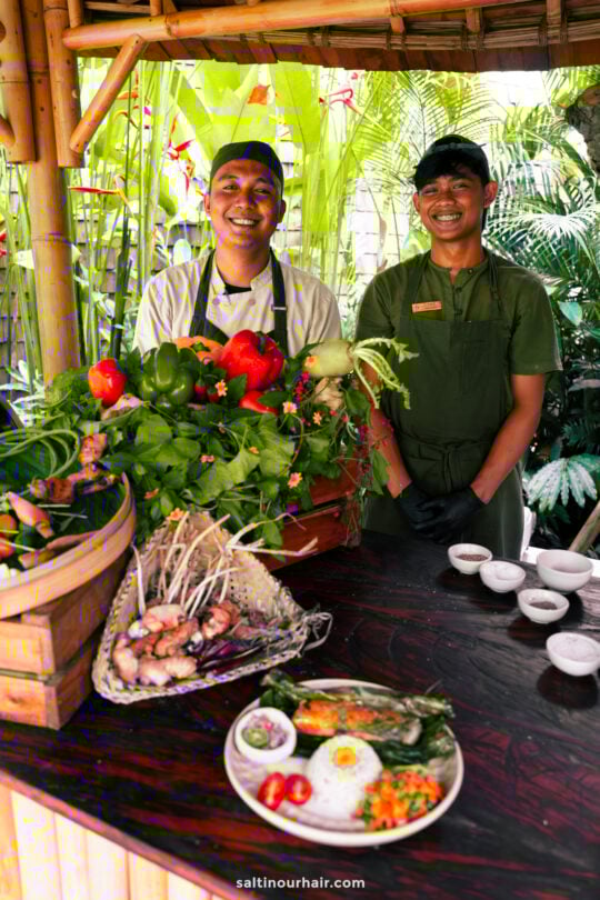 Two people in chef uniforms stand behind a wooden counter ready to give a cooking class at Bambootel Sawah view bali luxury hotel with fresh vegetables, herbs, spices, and a plated meal at a lush bambootel, surrounded by vibrant green plants.