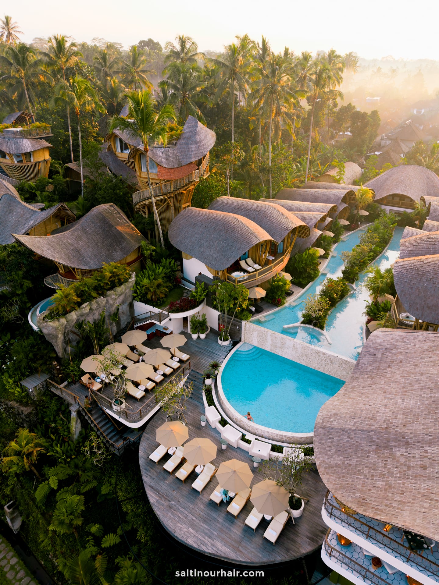 Aerial view of a tropical bambootel sawah view resort bali featuring curved, thatched-roof villas, swimming pools, sun loungers, and lush green palm trees.