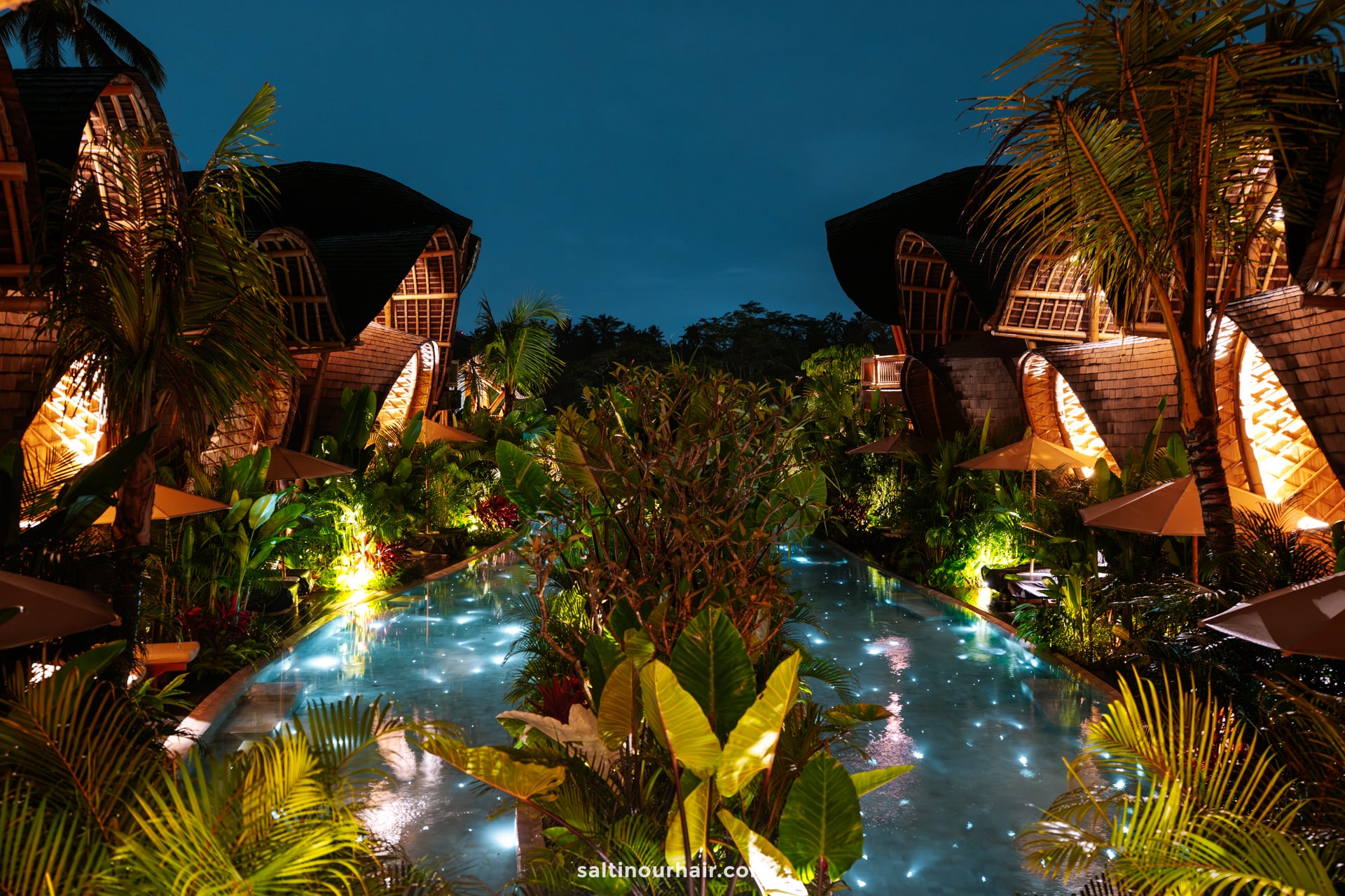 Nighttime view of a tropical bambootel resort bali featuring wooden villas, lush greenery, and a central illuminated swimming pool reflecting the surrounding lights.