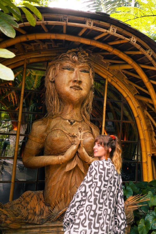 A woman stands in front of a large woven bamboo statue with hands in a prayer position, set against a lush bambootel structure surrounded by greenery.