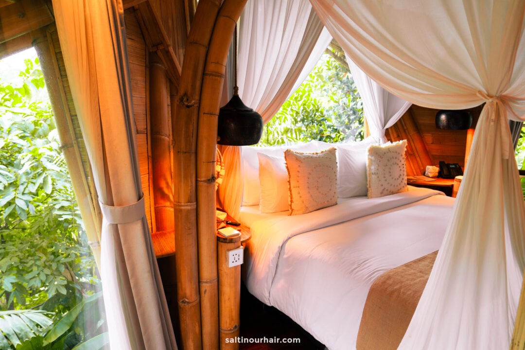 A cozy bambootel bedroom with a bamboo frame, white canopy curtains, a neatly made bed with white linens and pillows, and large windows overlooking lush green foliage of bali indonesia