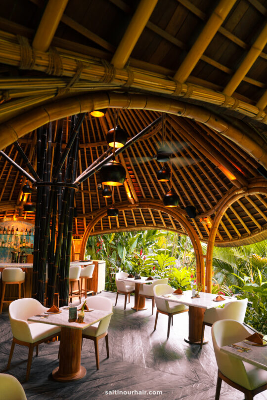 Modern tropical Maniq restaurant with bamboo architecture inspired by bambootel bali design, white chairs, wooden tables, and lush greenery visible through open arches. Warm lighting enhances the inviting atmosphere.