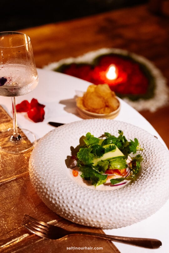 A white plate with a salad garnished with greens, a glass of clear drink, and a bowl of chips on a bambootel table set for dining, with a heart-shaped candle and rose petals in the background.