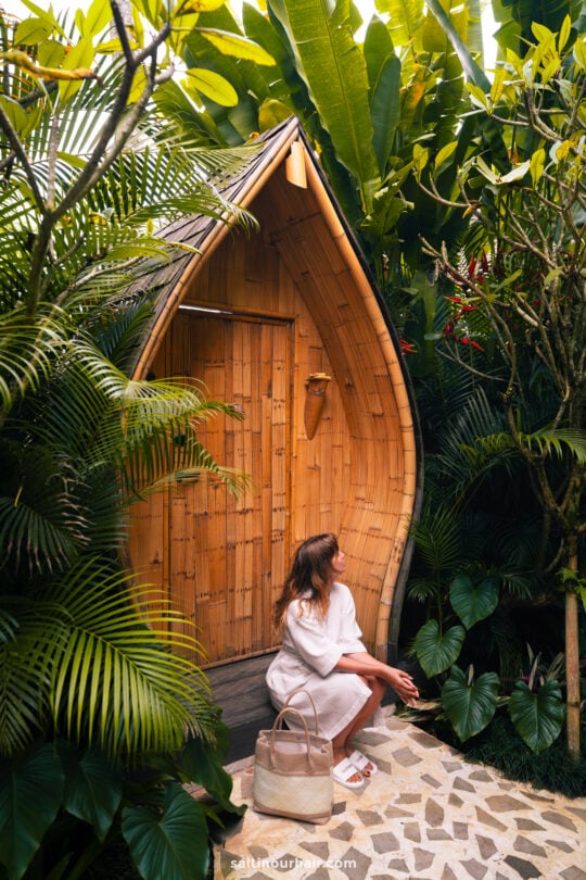 A woman in a white dress sits on the steps of a charming bambootel sawah view luxury hotel bali, surrounded by lush tropical plants, holding sunglasses and a beige bag.