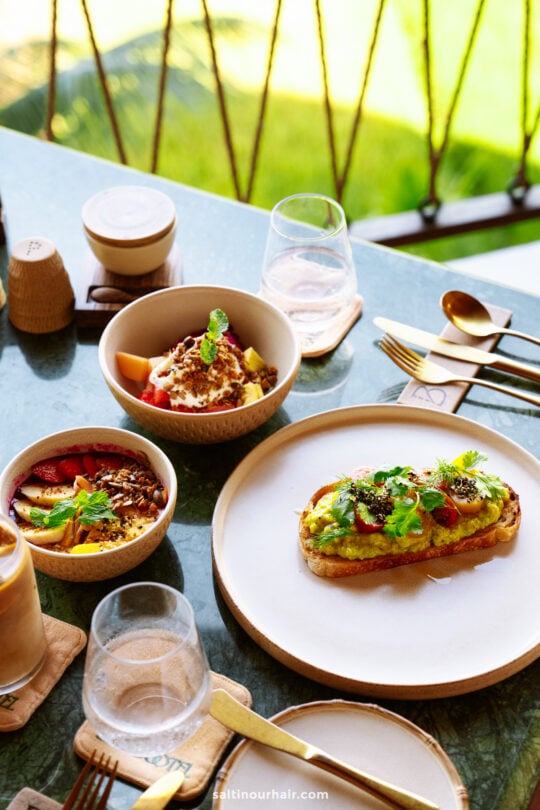 A table at Bambootel sawah view bali set with avocado toast, a smoothie bowl, a fruit bowl topped with granola, drinks, and utensils, with greenery visible in the background.