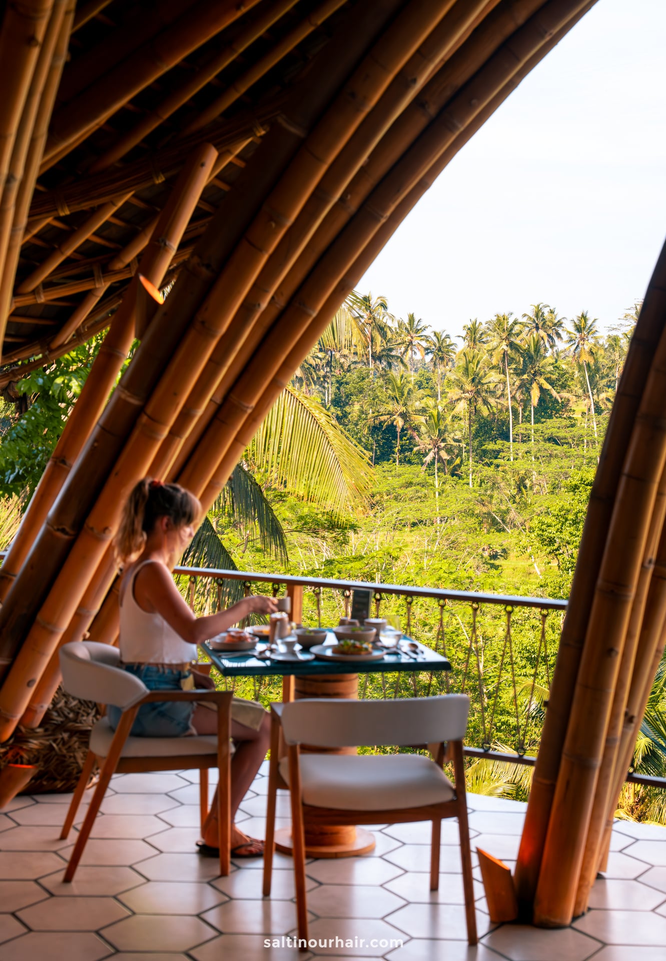 A woman sits alone at a table with food and drinks on a bamboo terrace at a bambootel luxury hotel, overlooking a lush tropical forest of bali indonesia