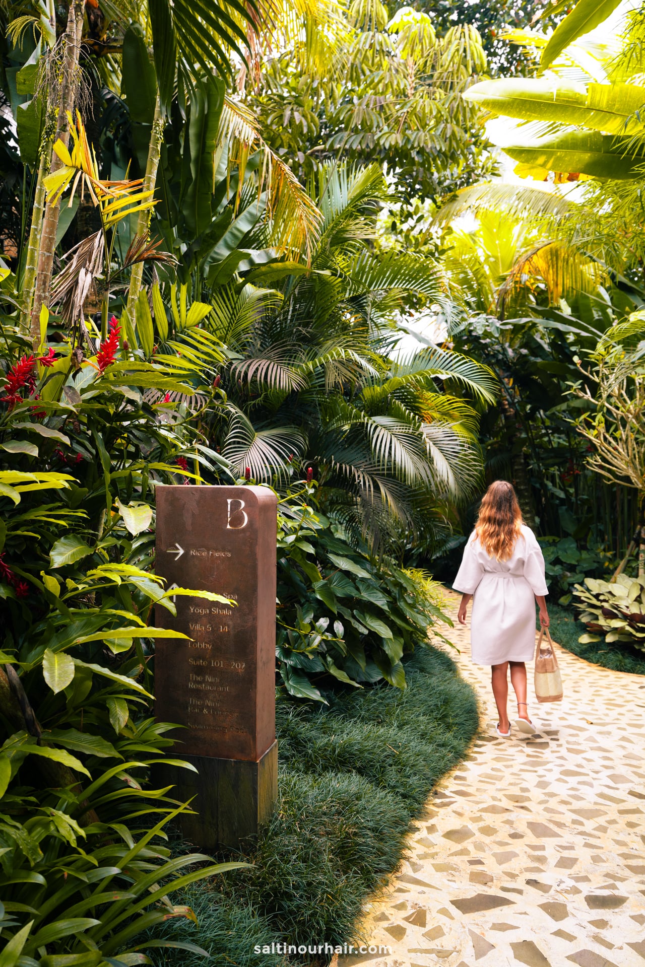 A woman with long hair in a white rope walks along a stone path surrounded by lush tropical plants at the bambootel sawah view hotel in bali, beside a brown signpost.