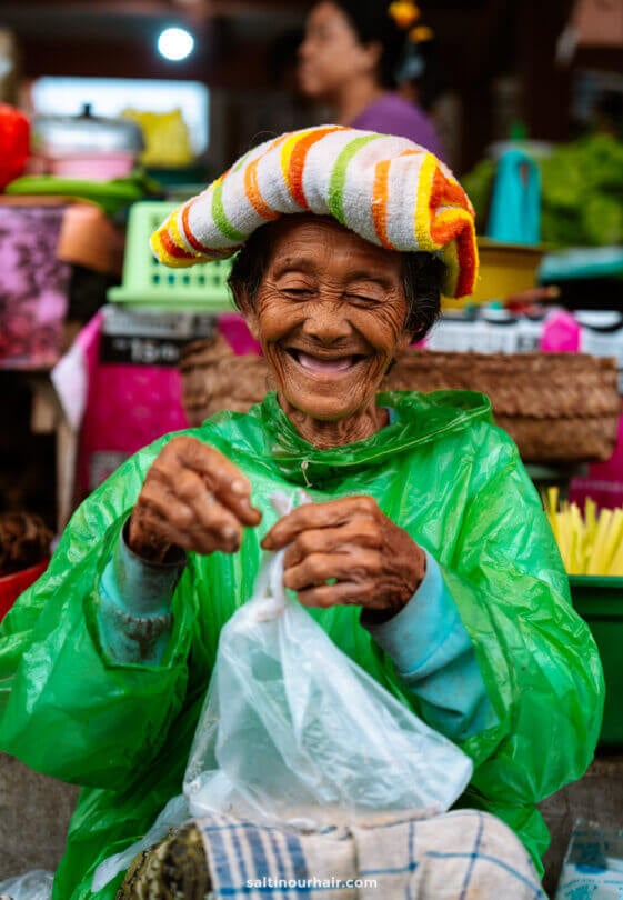 An elderly woman wearing a striped towel on her head and a green rain poncho smiles while handling a plastic bag at an outdoor market, capturing the vibrant spirit often experienced on a Bali cultural tour.