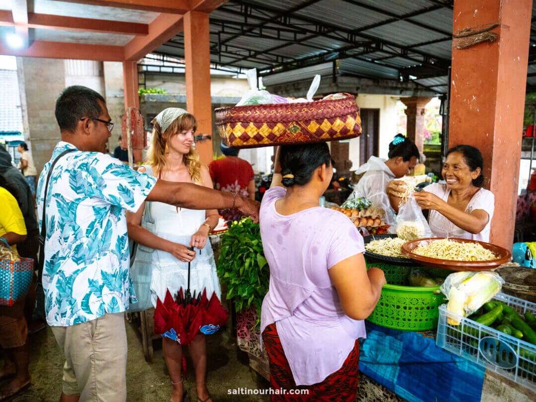 People shopping at a busy outdoor market during a Bali cultural tour; a woman carries a woven basket on her head while vendors sell produce and goods at their stalls.