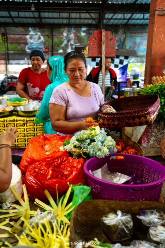 A woman arranges flowers at a busy market stall, surrounded by baskets and shoppers&mdash;a vibrant scene often experienced on a Bali cultural tour in a lively, covered, open-air market.