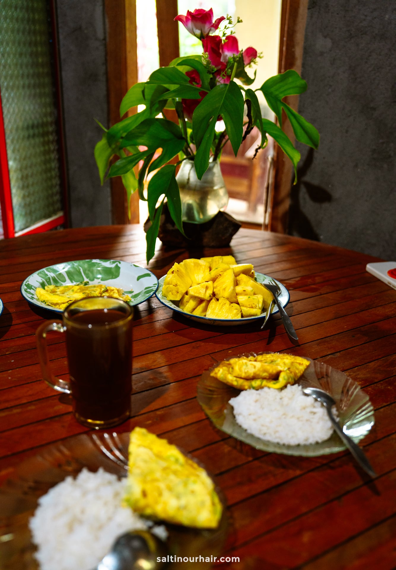 A wooden table set for a Bali cultural tour, featuring plates of rice and omelette, sliced pineapple, a mug of coffee, and a vase with red flowers.