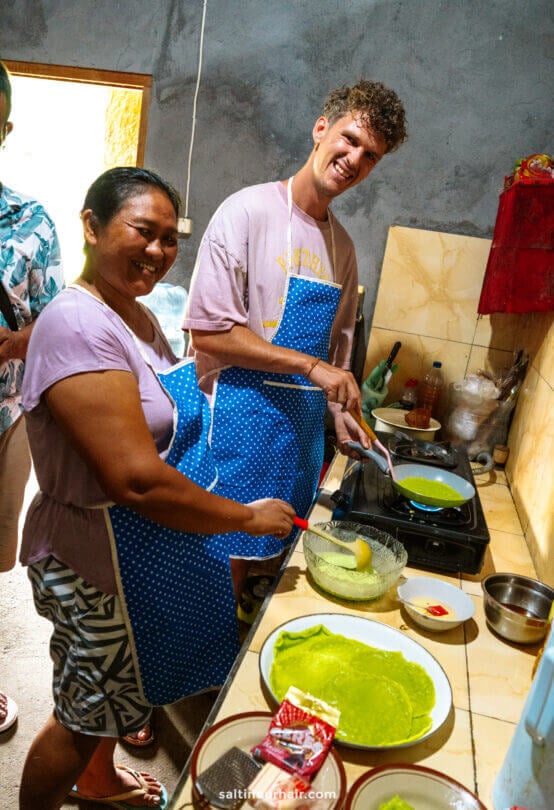 Two people wearing blue aprons smile while cooking green pancakes&mdash;reminiscent of treats from a Bali traditional cooking class&mdash;on a stove in a kitchen. Plates, utensils, and ingredients are spread across the counter.