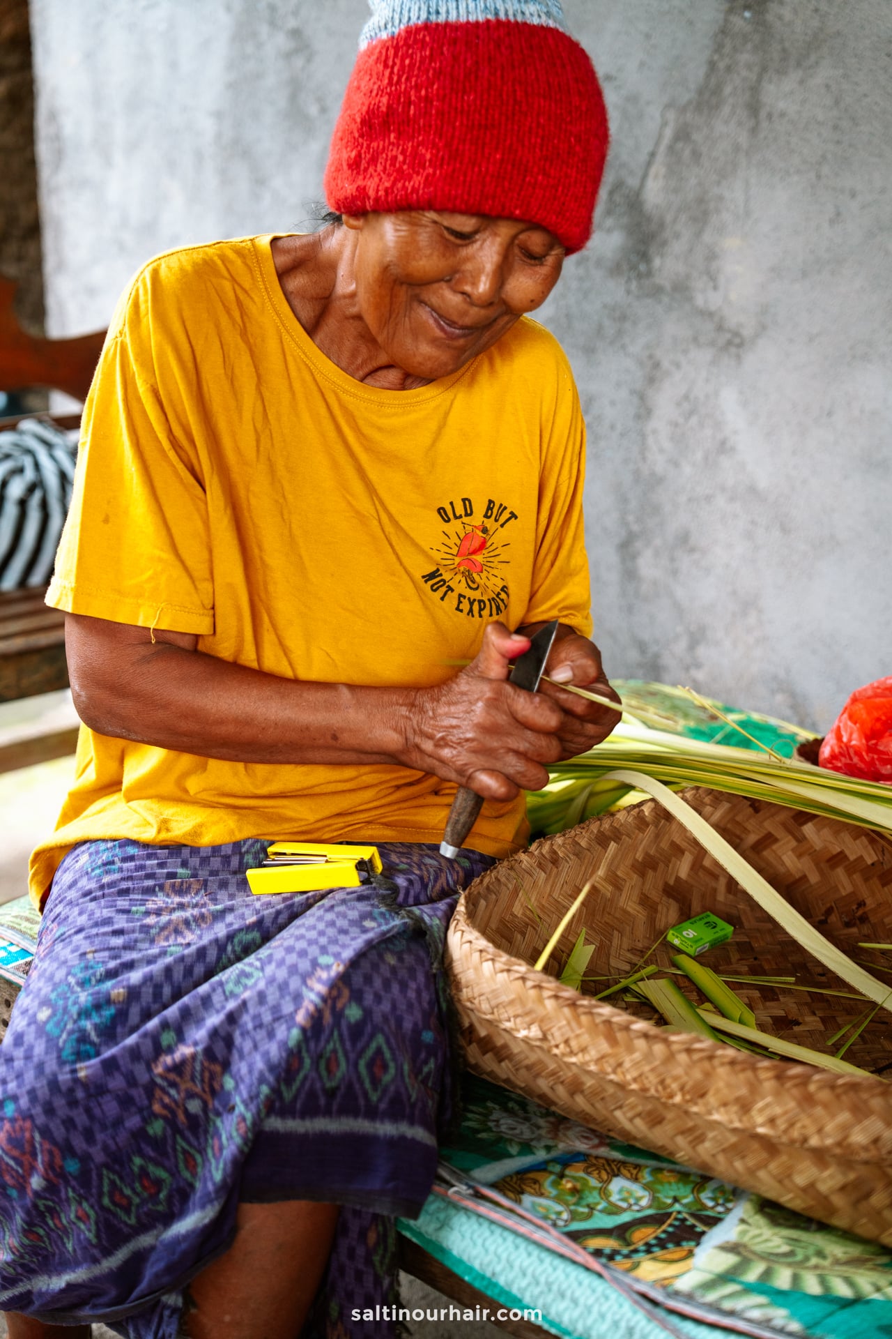 An elderly woman in a yellow shirt and red beanie weaves palm leaves by hand, sitting on a mat with a woven basket beside her&mdash;a scene commonly experienced on a Bali cultural tour.