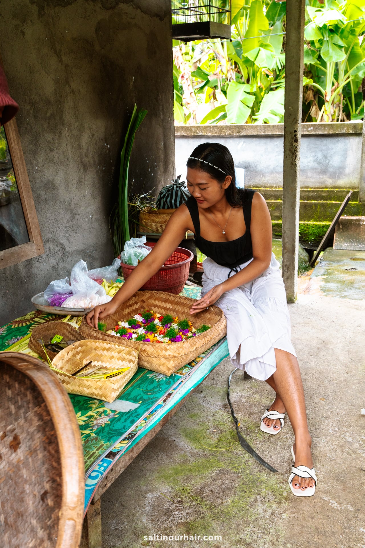 A woman sits on a bench preparing traditional balinese offerings with colorful flowers and leaves, surrounded by baskets and bags&mdash;an authentic scene from this Bali local cultural tour.