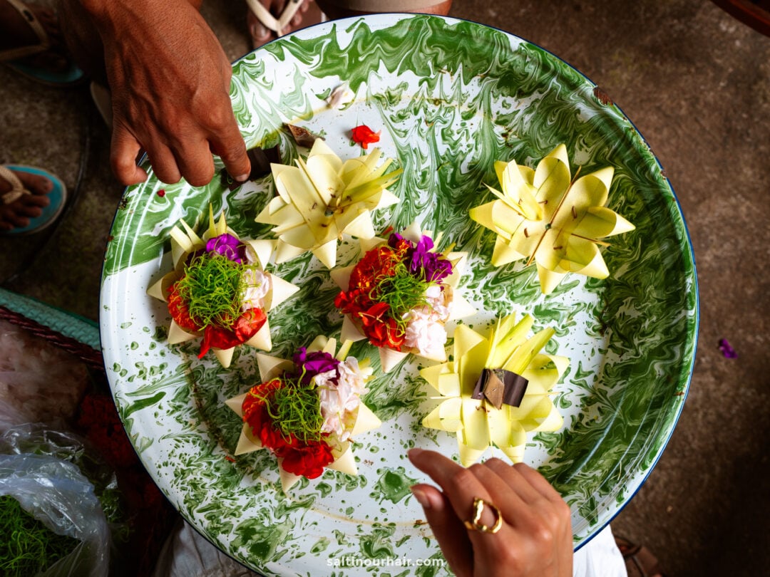 A green and white marbled tray holds five decorative floral arrangements made from yellow leaves, red and purple flowers, and small brown pieces&mdash;an artful scene often admired on a Bali traditional tour. Two hands are thoughtfully arranging the items.