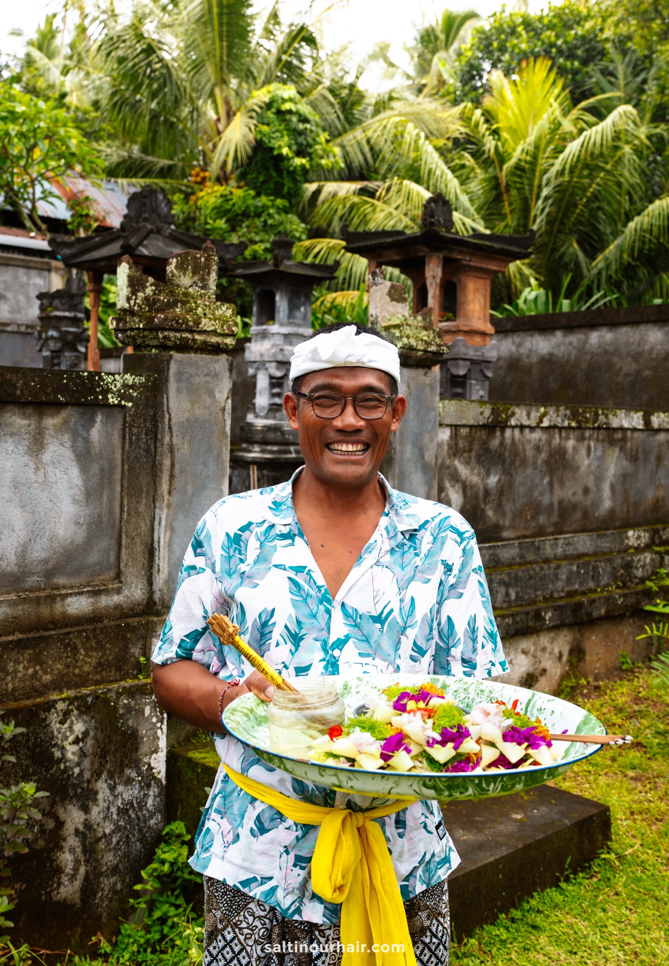 A man wearing glasses and a patterned shirt holds a large platter of food with flowers, standing outdoors in front of a stone wall and tropical greenery during a Bali traditional tour.