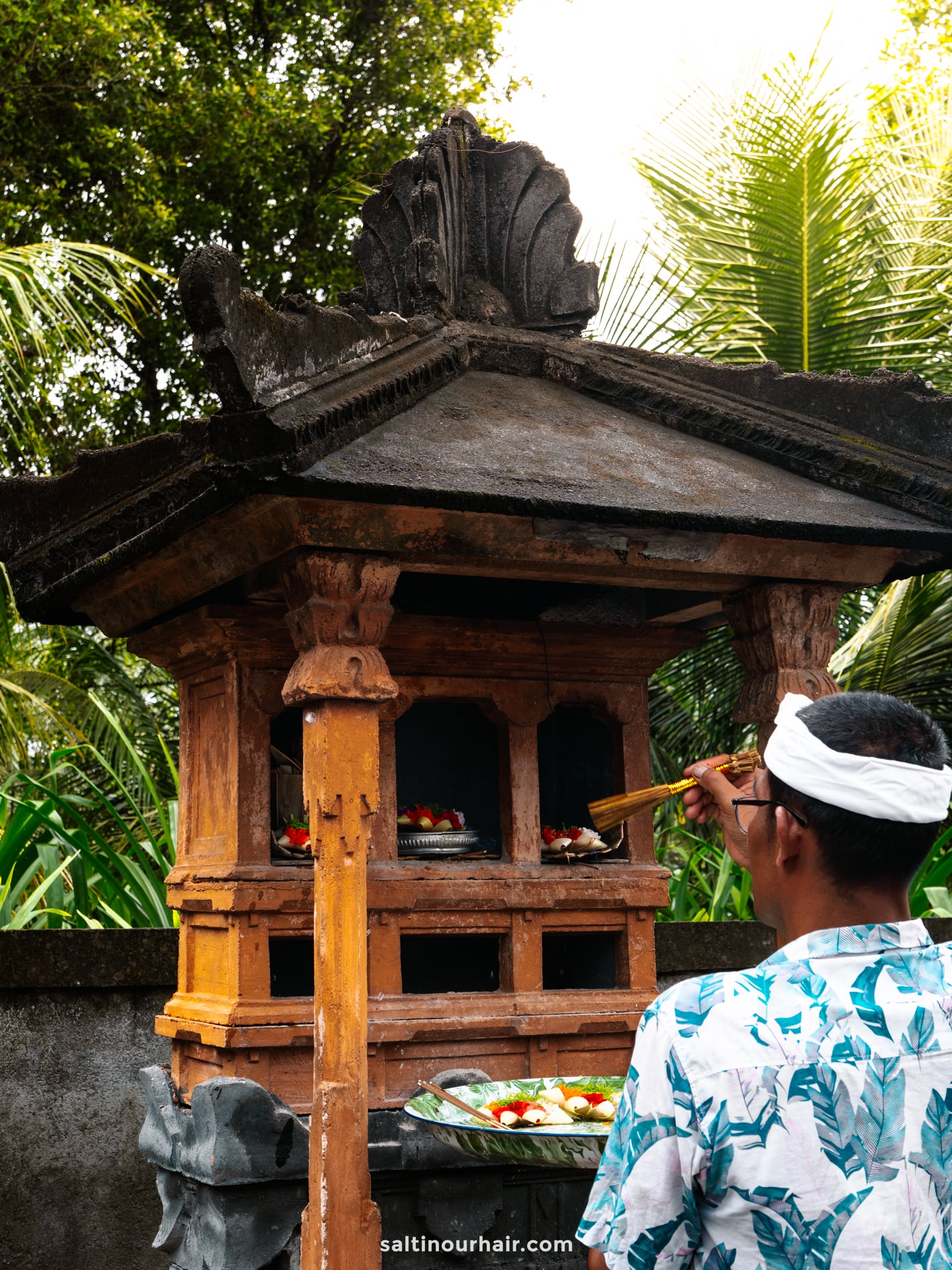 A man in a patterned shirt and headband offers incense at a traditional Balinese shrine, surrounded by greenery and fruit offerings&mdash;a captivating moment from a Bali cultural tour.