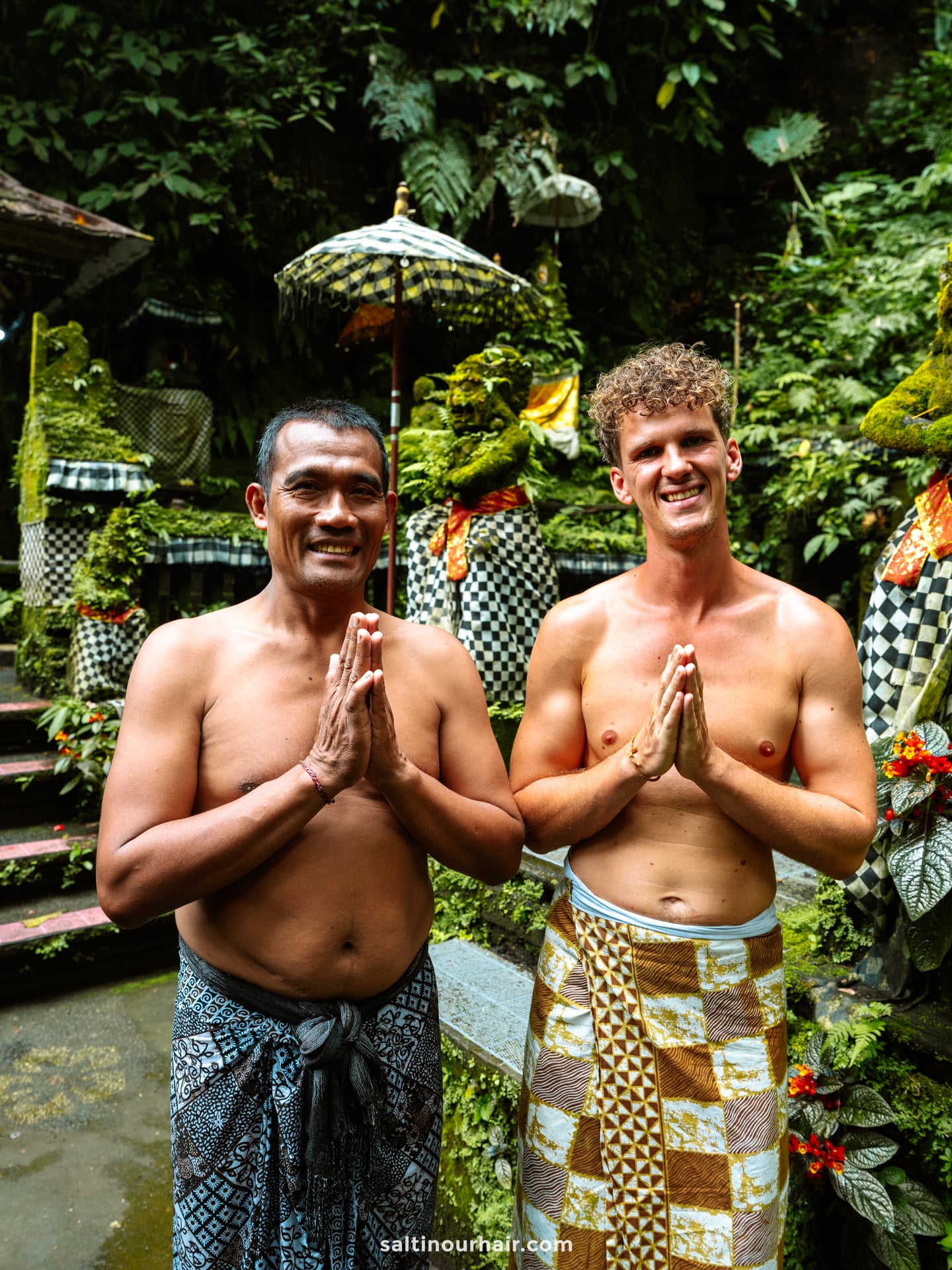 Two men wearing patterned sarongs stand side by side outdoors with palms pressed together in greeting, surrounded by lush greenery and traditional Balinese decorations&mdash;an authentic scene from a Bali cultural tour.