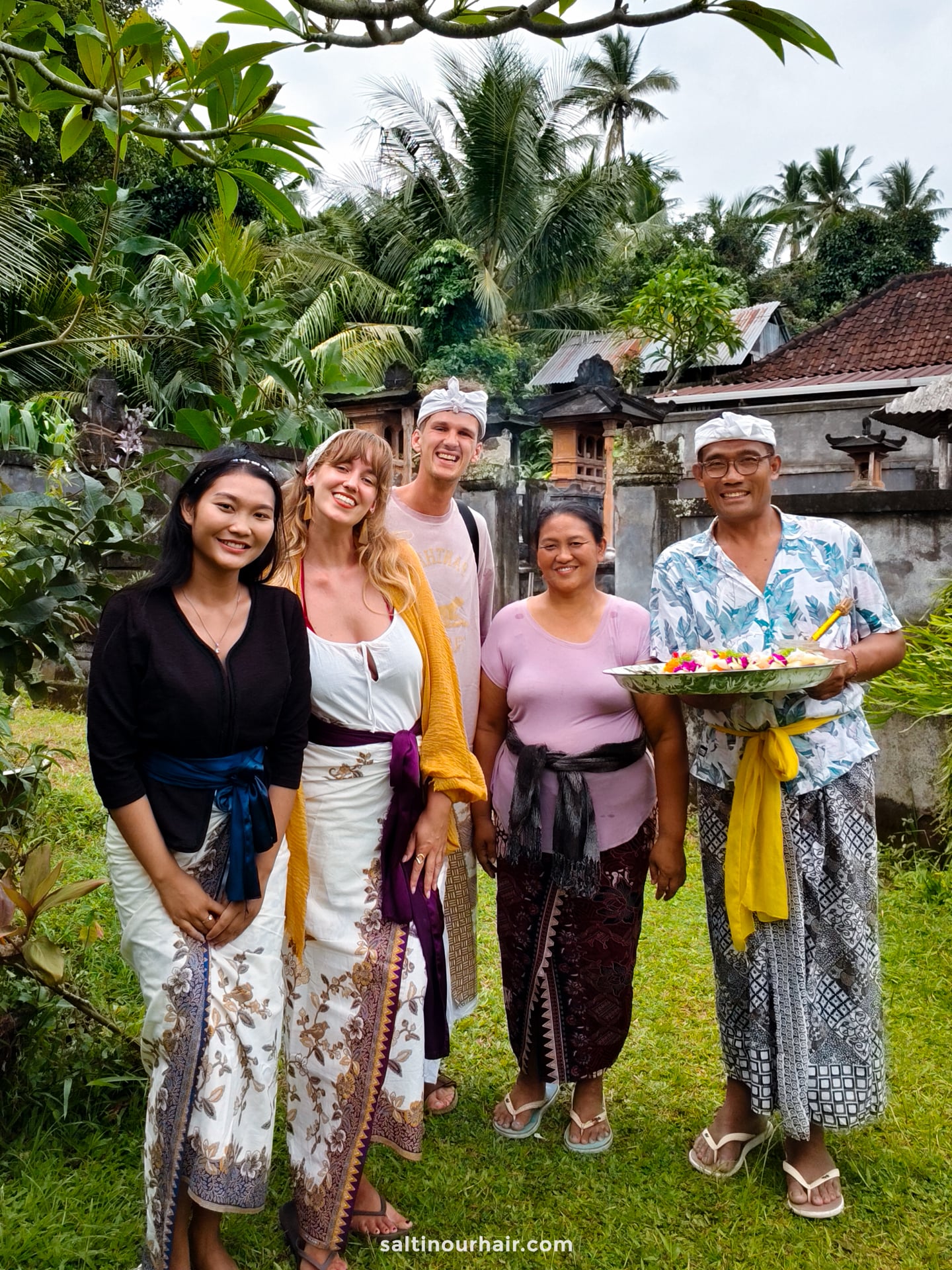 Five people stand outdoors in traditional Balinese attire, smiling for the camera during a Bali cultural tour in a lush garden with tropical plants and stone structures in the background.