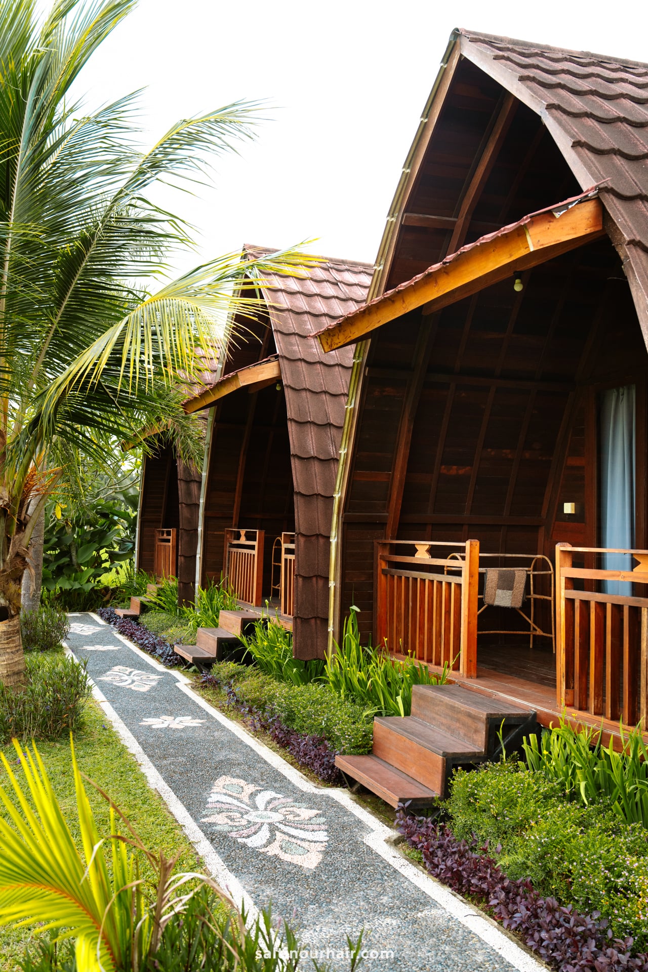 Three wooden cabins with arched roofs and small porches stand in a row, surrounded by tropical plants and a stone pathway&mdash;an inviting retreat after a day on your Bali cultural tour.