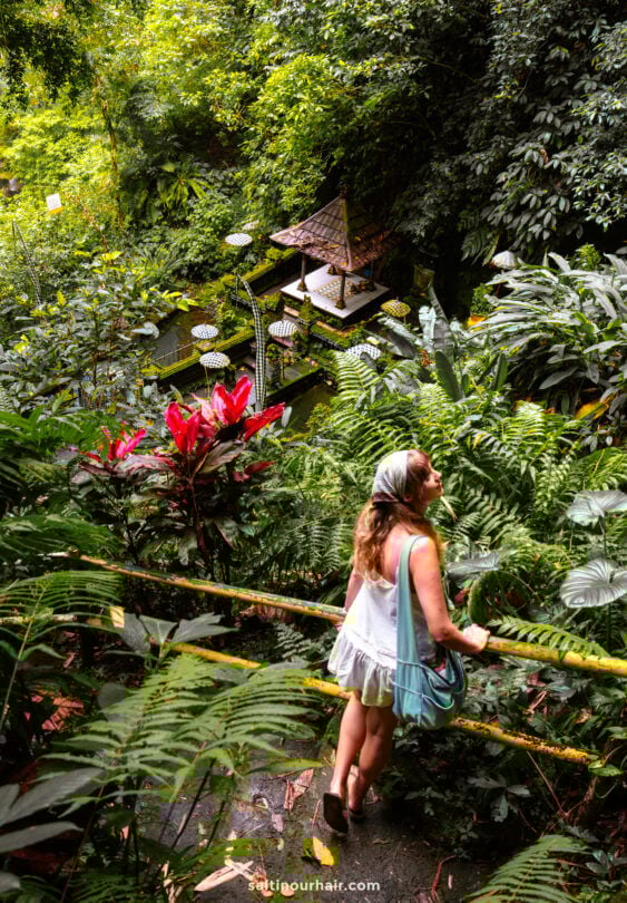 A woman in a white dress stands on a path surrounded by lush tropical plants, overlooking a small pavilion in a verdant garden&mdash;capturing the serene elegance of a Bali cultural tour.