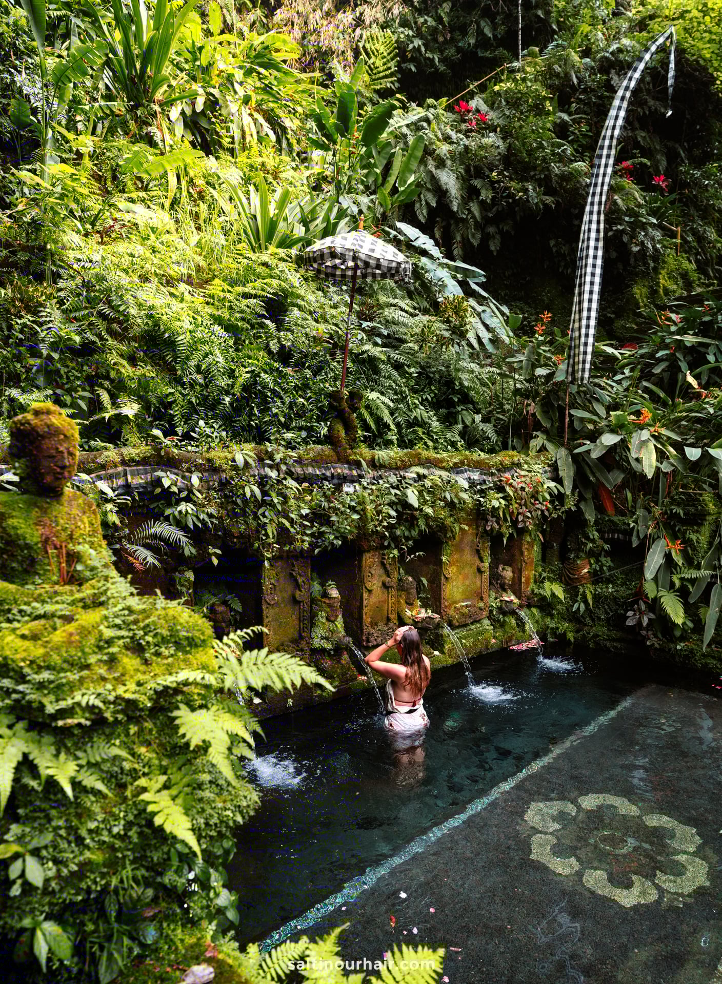 A woman stands under water spouts in a stone pool, surrounded by lush, green tropical plants and mossy walls&mdash;a serene moment on her Bali cultural tour in a jungle setting.