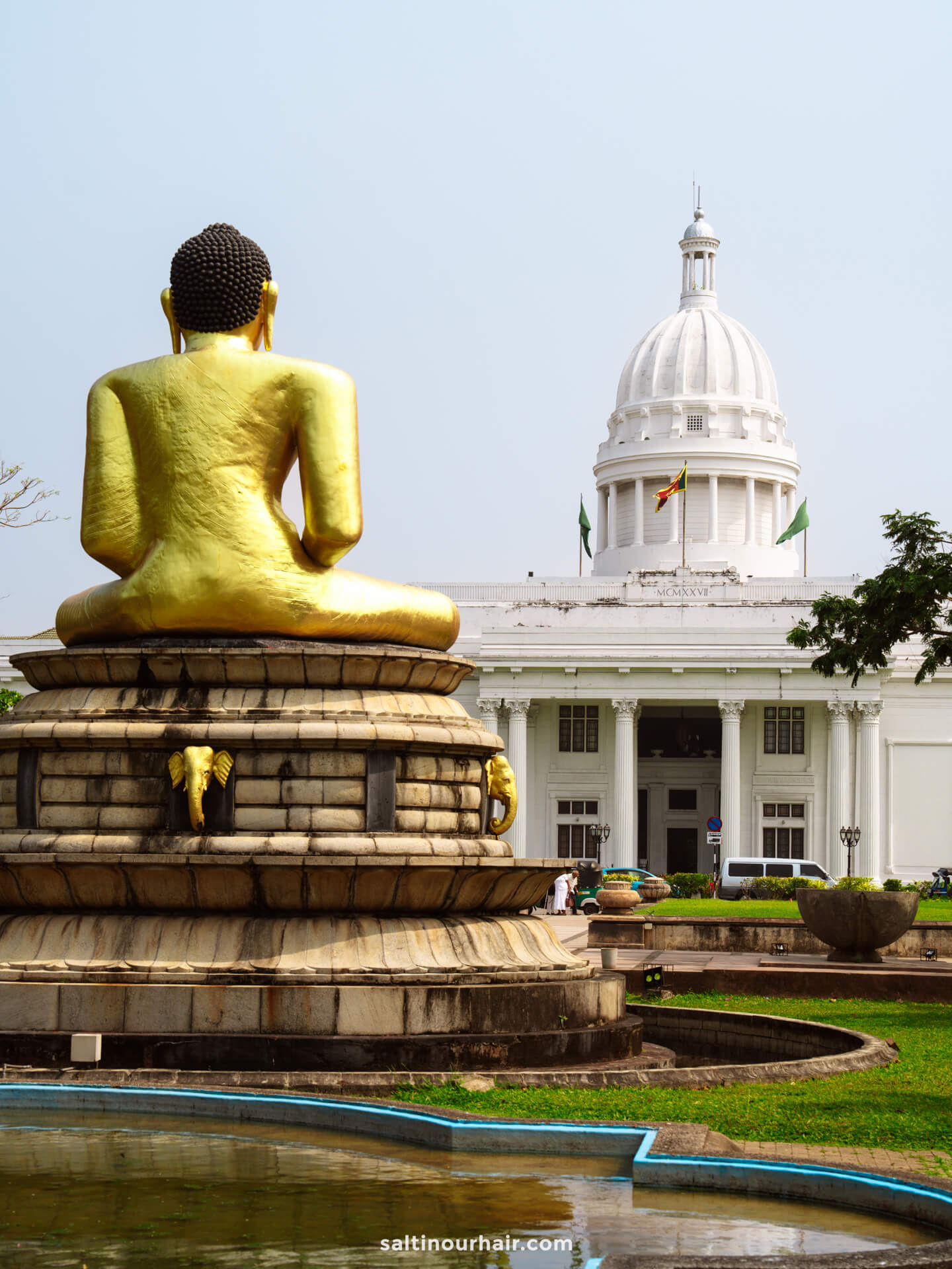 A large golden Buddha statue sits before a white domed government building with columns and a flag on top, surrounded by grass and a fountain&mdash;one of the must-see things to do Colombo offers for history and culture lovers.