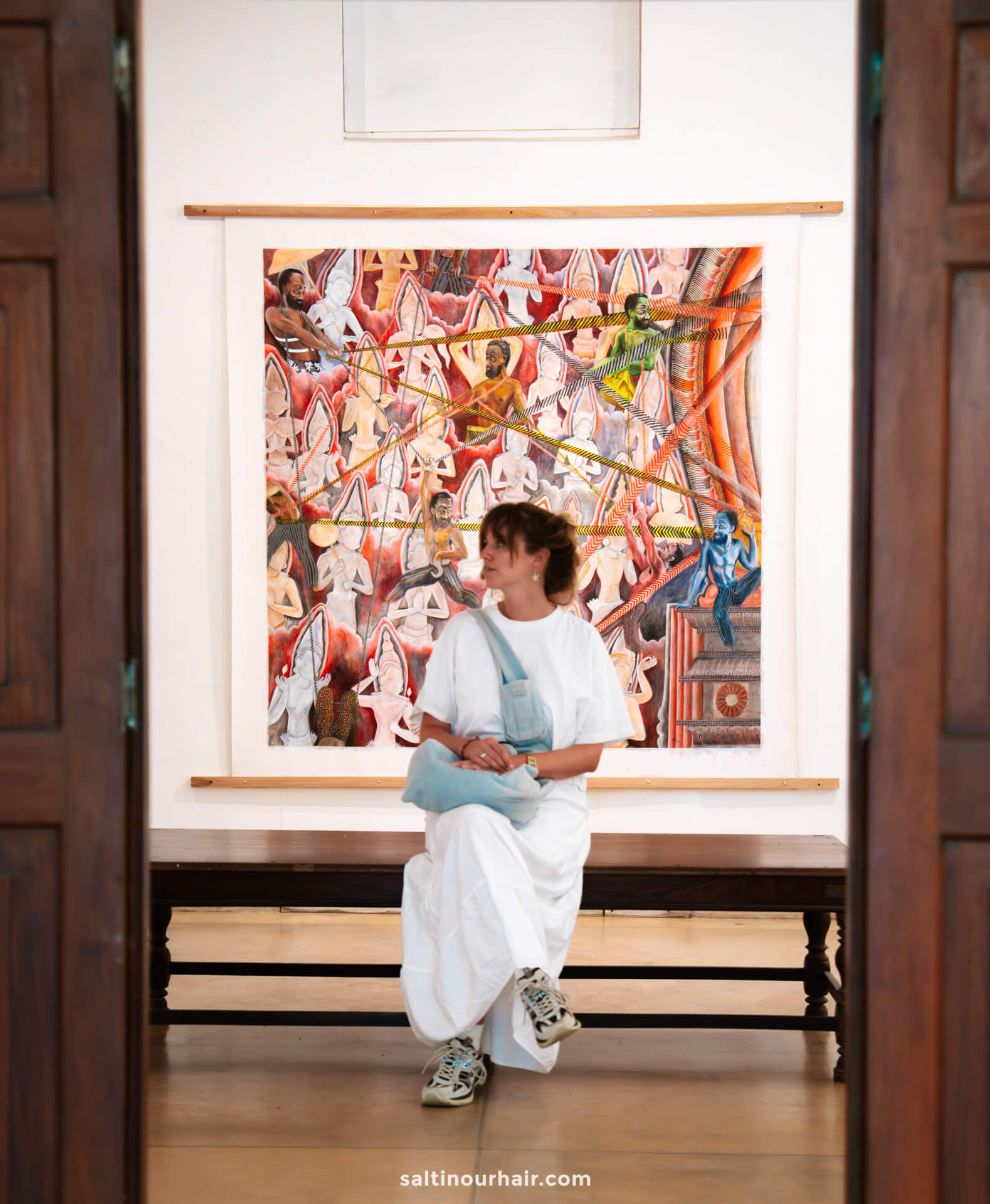 A woman in a white dress sits on a bench in front of a colorful, abstract painting displayed on a gallery wall&mdash;one of the must-see things to do Colombo offers for art enthusiasts.