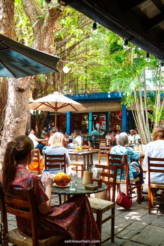 People sitting at wooden tables under large trees in an outdoor caf&eacute;, with umbrellas providing shade and lush greenery all around&mdash;a perfect spot to relax and experience one of the best things to do Colombo has to offer.