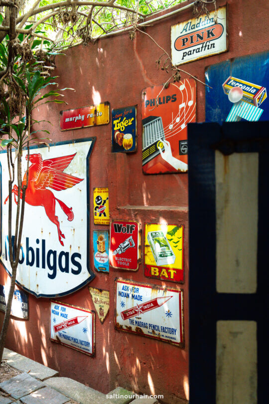 Vintage metal advertisement signs, including Mobilgas and various product ads, are mounted on a sunlit red brick wall, partially shaded by nearby plants&mdash;evoking retro charm reminiscent of unique things to do Colombo offers.