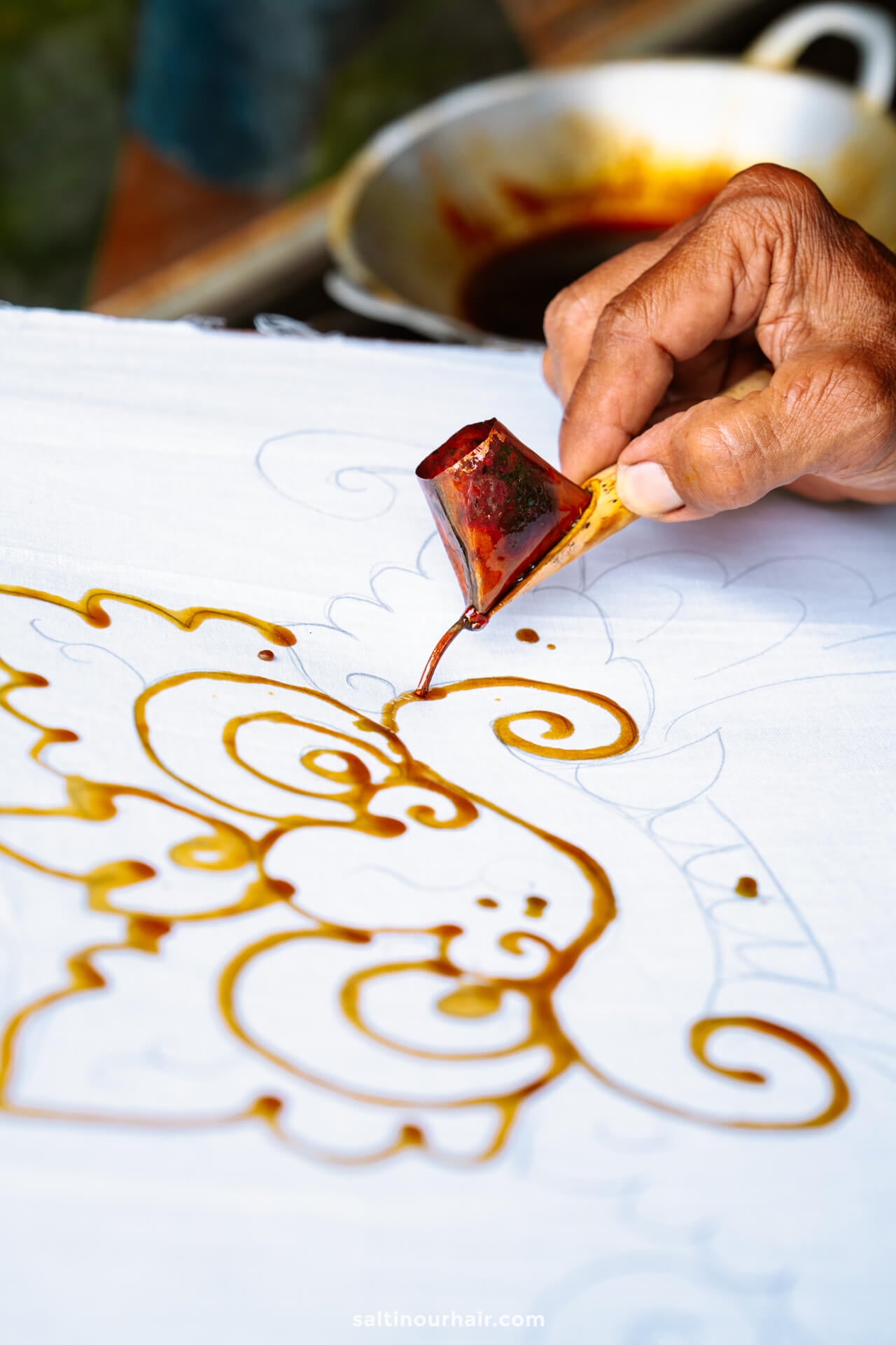 A hand applies hot wax in intricate patterns onto white fabric during a batik painting workshop, with a pot of wax visible in the background.