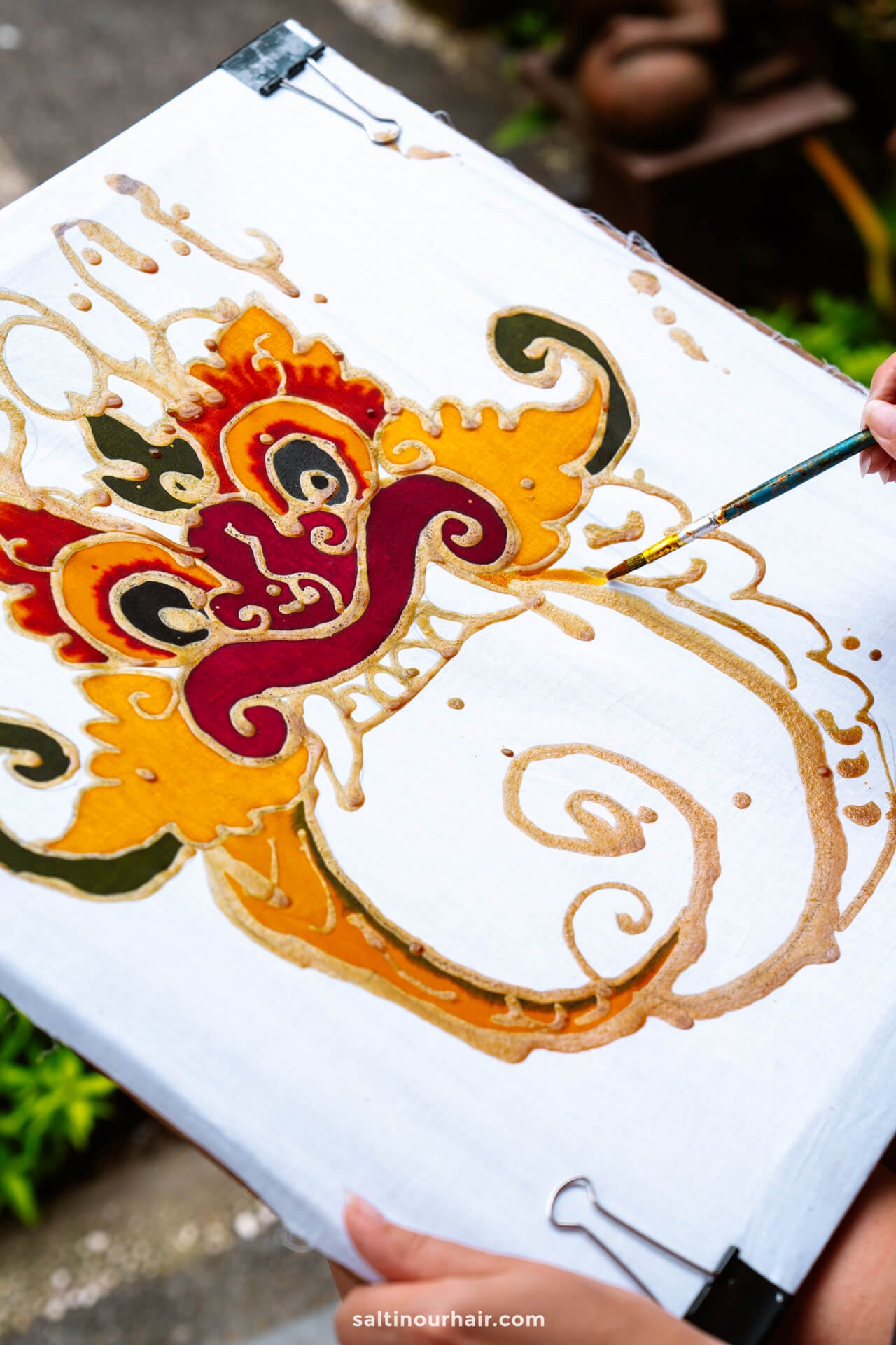 A person participates in a batik painting workshop in bali, creating a traditional, ornate design in red, yellow, and gold on a white cloth stretched on a frame outdoors.