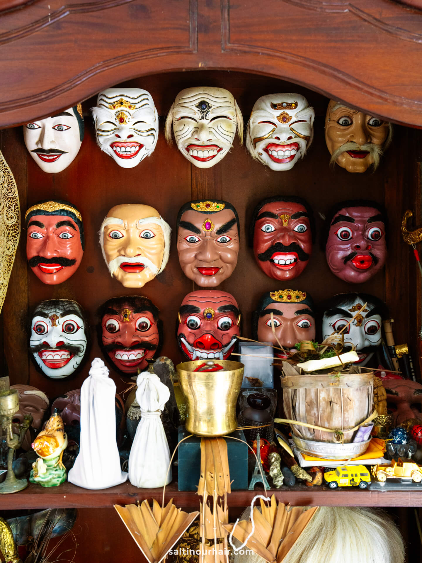 A wooden shelf displays various colorful traditional balinese masks with exaggerated expressions, along with assorted objects such as candles, a brass bowl, small figurines, and supplies from a local batik painting workshop.