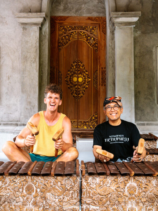 Two men sit side by side playing wooden xylophones called Gamelan in front of an ornately carved door; one wears a yellow tank top, the other glasses and a traditional balinese headscarf