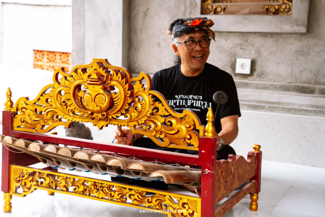 A man wearing glasses and traditional headgear plays a Balinese gamelan instrument with ornate yellow and red carvings indoors, beside a vibrant batik painting workshop in progress.