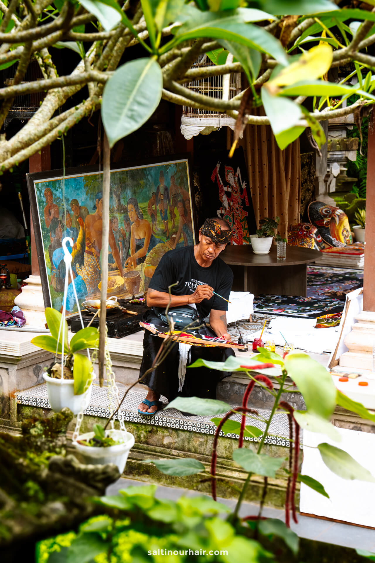 A man sits outdoors under a tree, painting on fabric with brushes during a batik painting workshop in bali. Colorful finished artwork and painting supplies surround him.