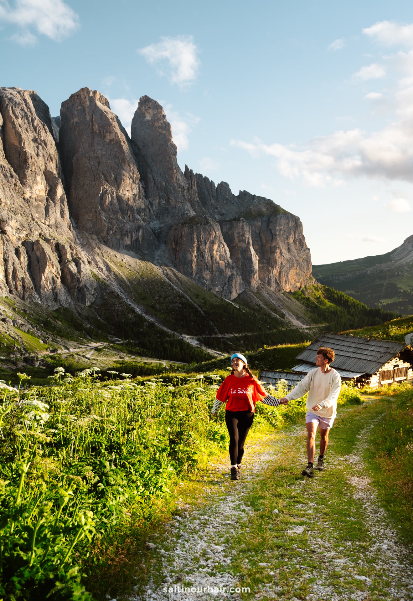 Two people walk on a mountain path surrounded by greenery, with rocky peaks and a wooden cabin in the background&mdash;an inspiring scene for your Dolomites itinerary.