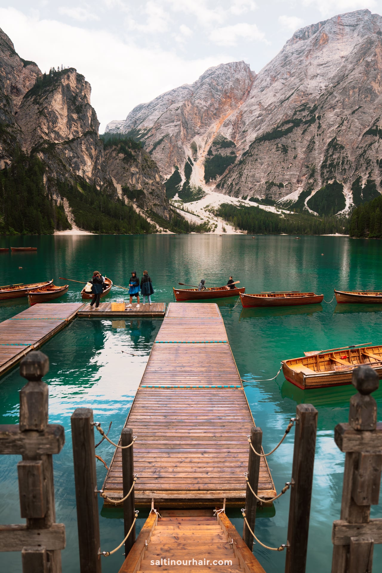 A wooden dock extends into a turquoise lake with several wooden boats, surrounded by mountains and forested slopes under a cloudy sky&mdash;an idyllic spot to include in your Dolomites itinerary.