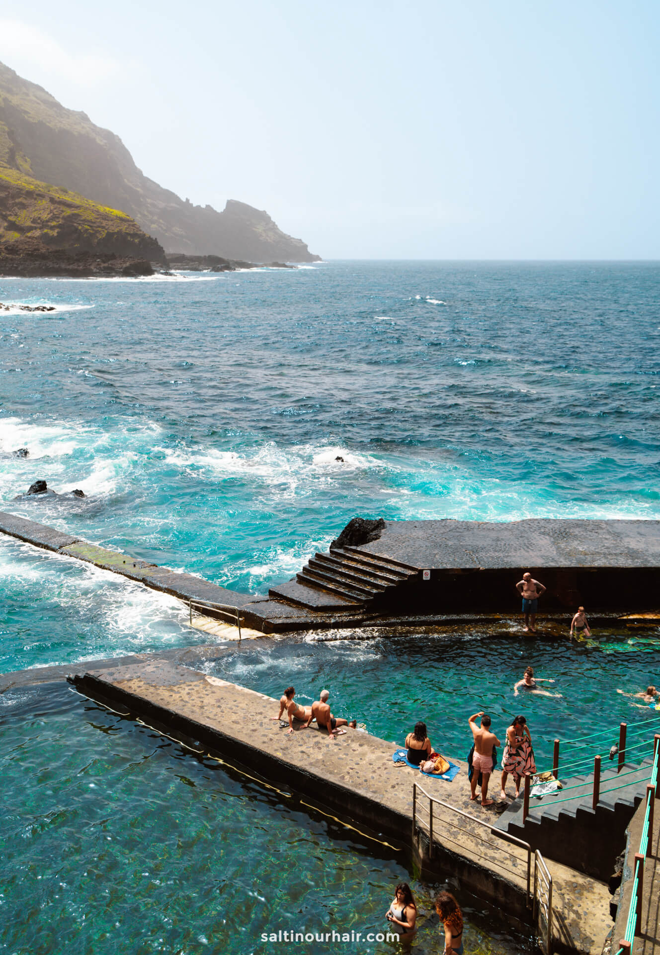 People swim and relax in a natural ocean pool beside rocky cliffs on La Palma, with waves crashing and the sea stretching toward a misty Canary Islands horizon.