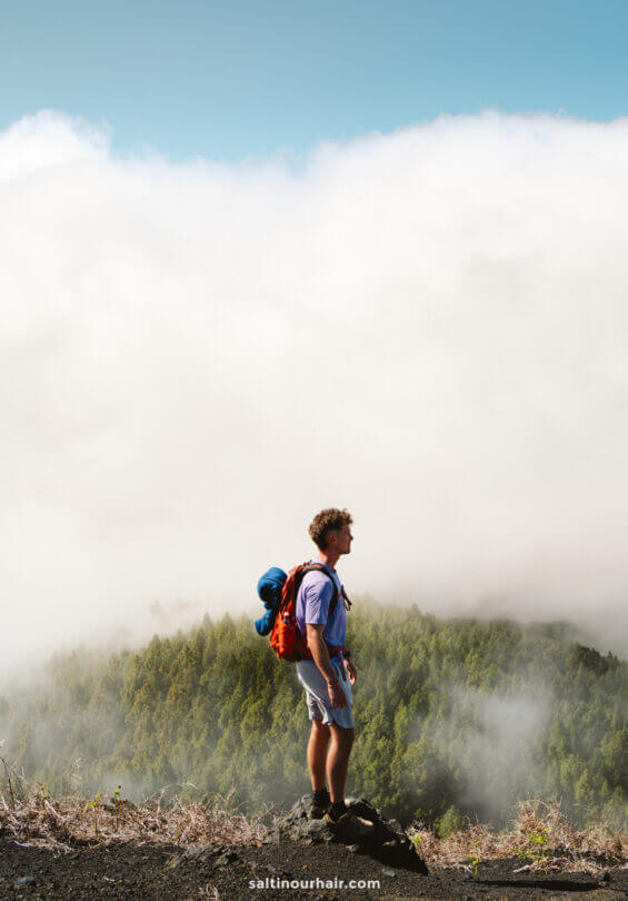 A man with a red backpack stands on a rocky ledge overlooking La Palma’s forested landscape, clouds drifting beneath a bright blue sky—a perfect scene for any Canary Islands travel guide.