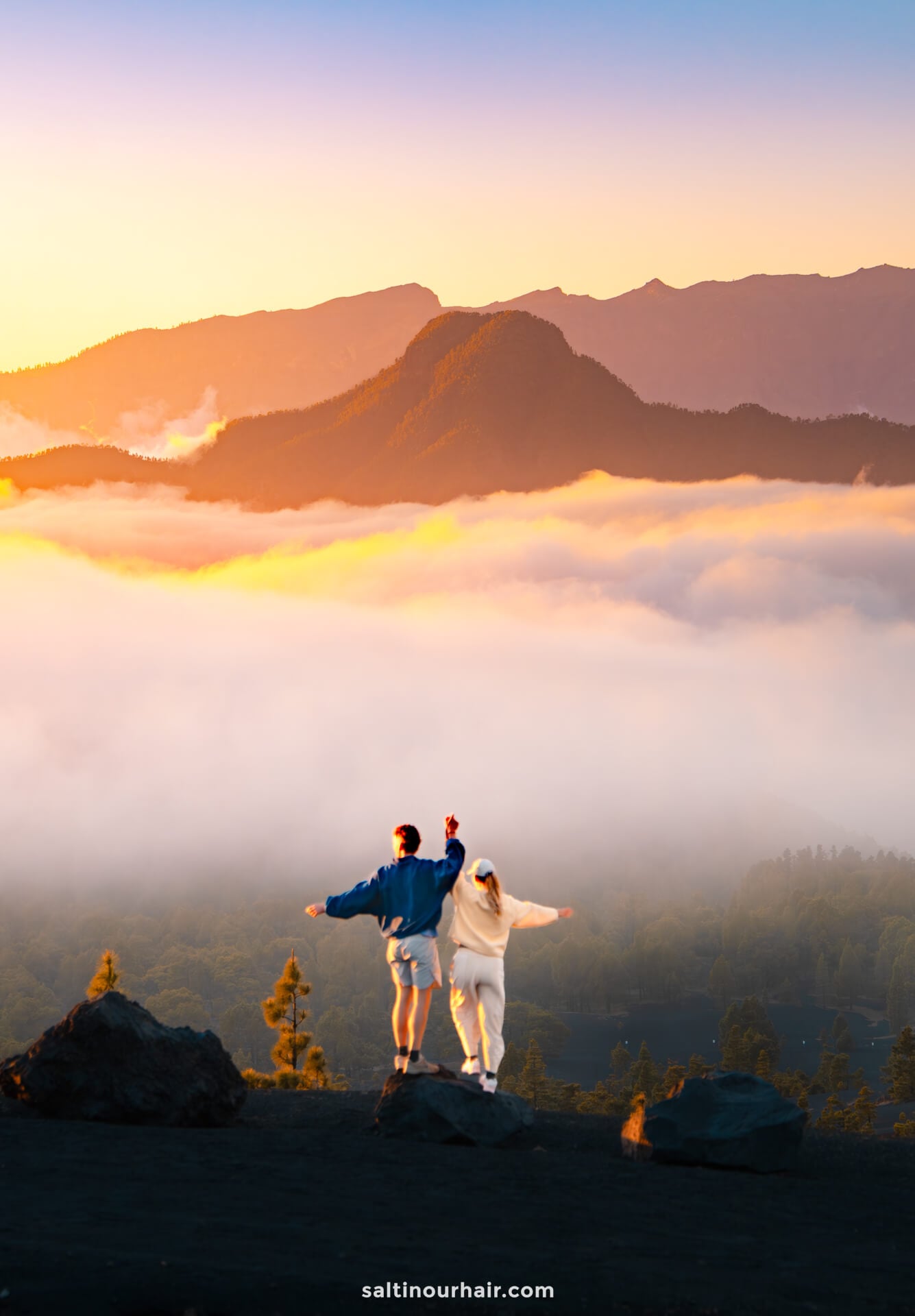 Two people stand on rocks overlooking a foggy valley in La Palma, Canary Islands, with mountains in the background during sunrise, wearing casual clothes and facing the view—an inspiring moment from a Travel Guide.