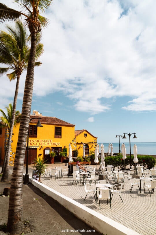 Outdoor restaurant patio with empty white tables and chairs, palm trees, and a yellow building near the ocean under a partly cloudy sky—an inviting La Palma scene from a Canary Islands travel guide.