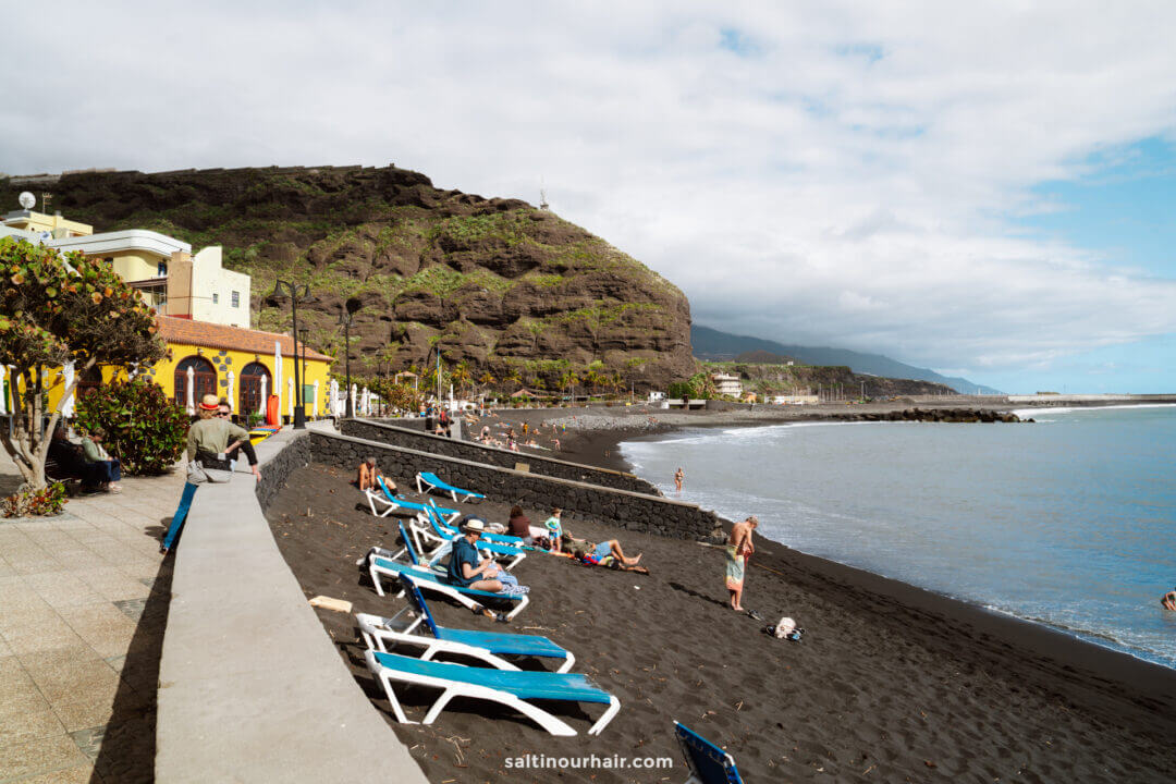 People relax on blue lounge chairs on a black sand beach in La Palma, bordered by a stone wall, with cliffs and colorful buildings in the background under a partly cloudy sky—a perfect scene from the Canary Islands Travel Guide.