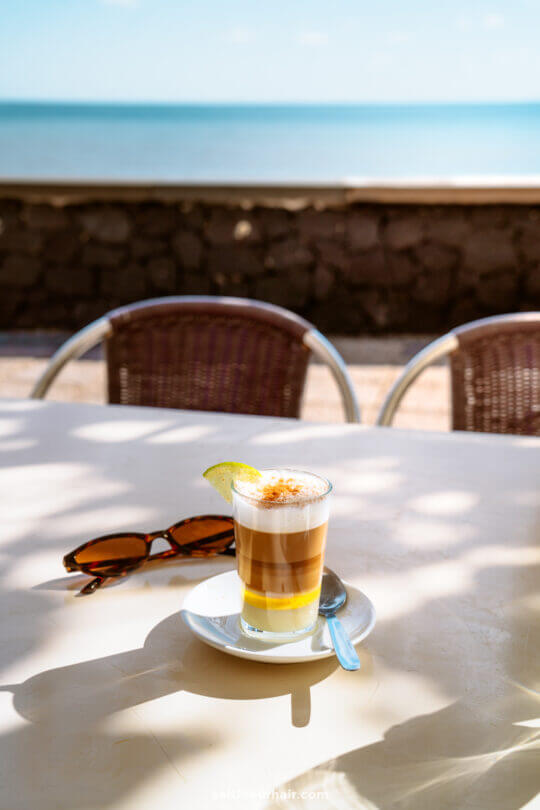 A layered coffee with a lemon slice sits on a saucer with a spoon on a sunlit outdoor table in La Palma, next to sunglasses, with the ocean and a stone wall in the background—a perfect scene for any Canary Islands travel guide.