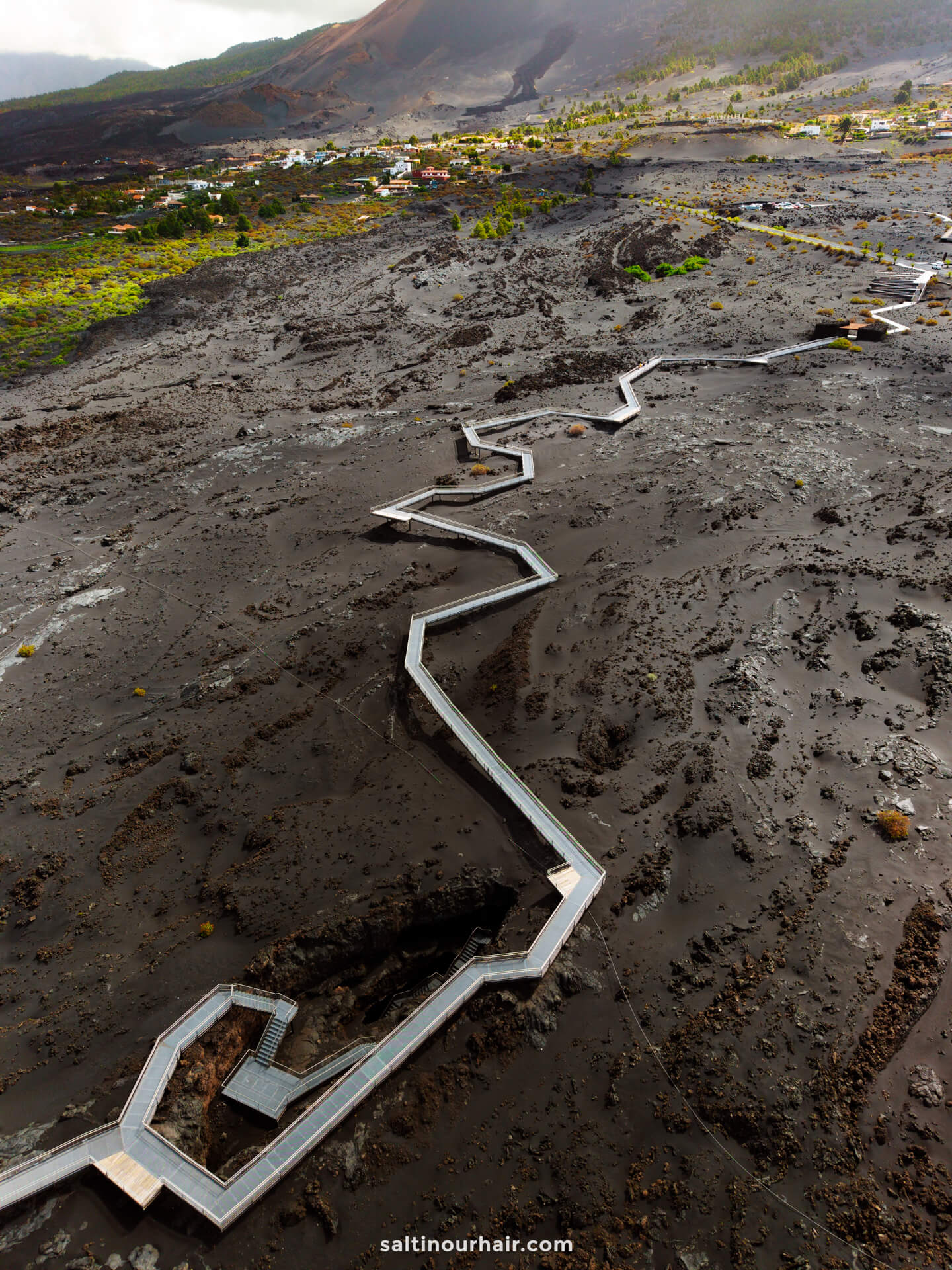 A zigzag wooden walkway crosses a barren, volcanic ash landscape on La Palma, with small houses and green vegetation in the background—an iconic sight in this Canary Islands travel guide.