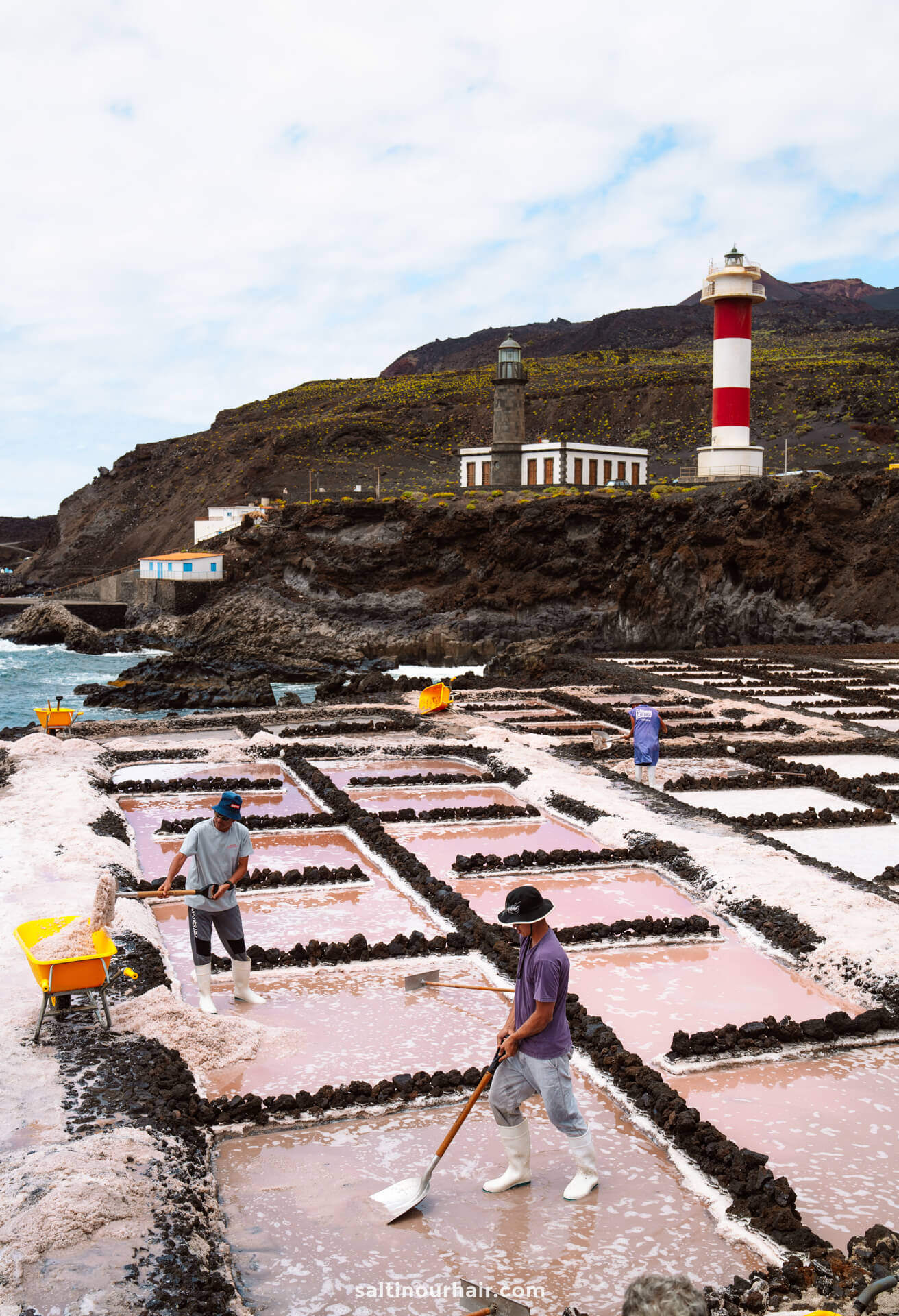 Workers harvest salt from rectangular salt pans near the coast of La Palma, with a lighthouse and buildings on a rocky hill in the background—a striking scene in the Canary Islands worthy of any travel guide.
