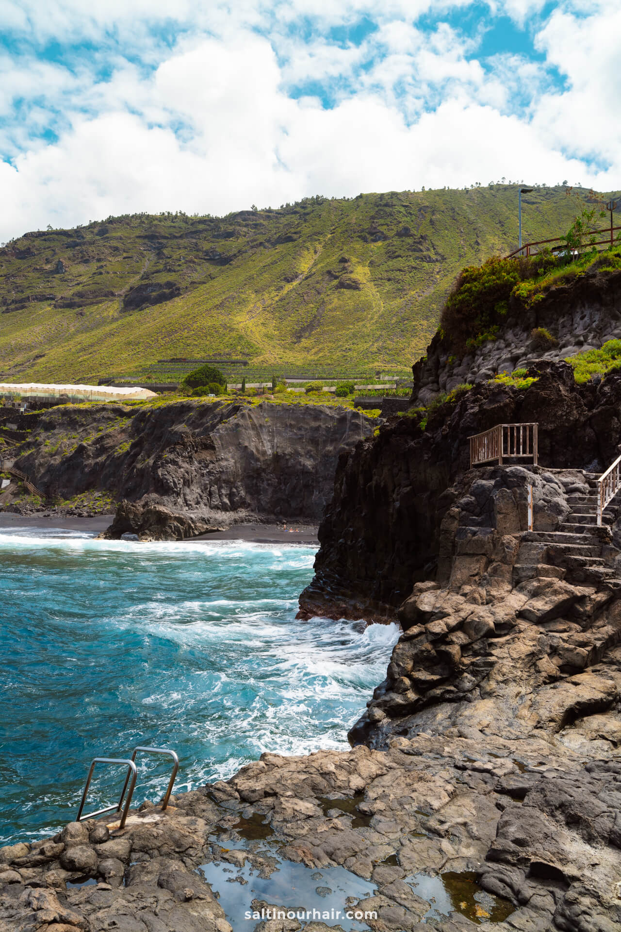 Rocky coastline with metal stairs leading into the ocean on La Palma, waves crashing against the rocks, and green hills in the background under a partly cloudy sky—an iconic scene from any Canary Islands travel guide.