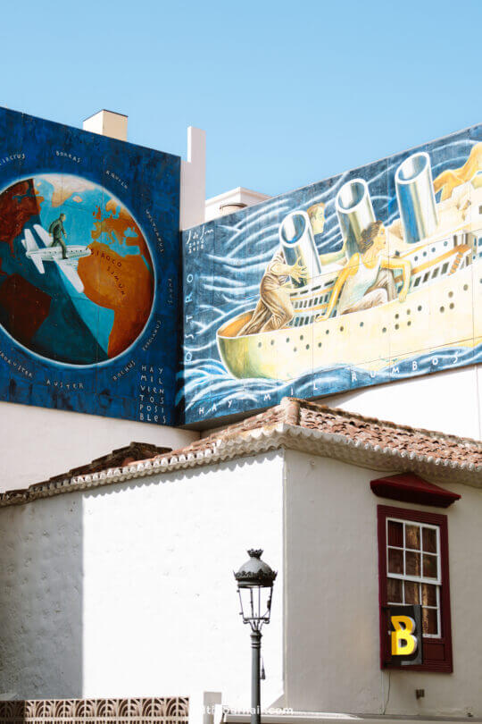 Two colorful murals on a building wall in La Palma: one shows an airplane over a globe, the other depicts a large ship on water. A white building with a red window and a yellow B is in the foreground—perfect for any Canary Islands travel guide.