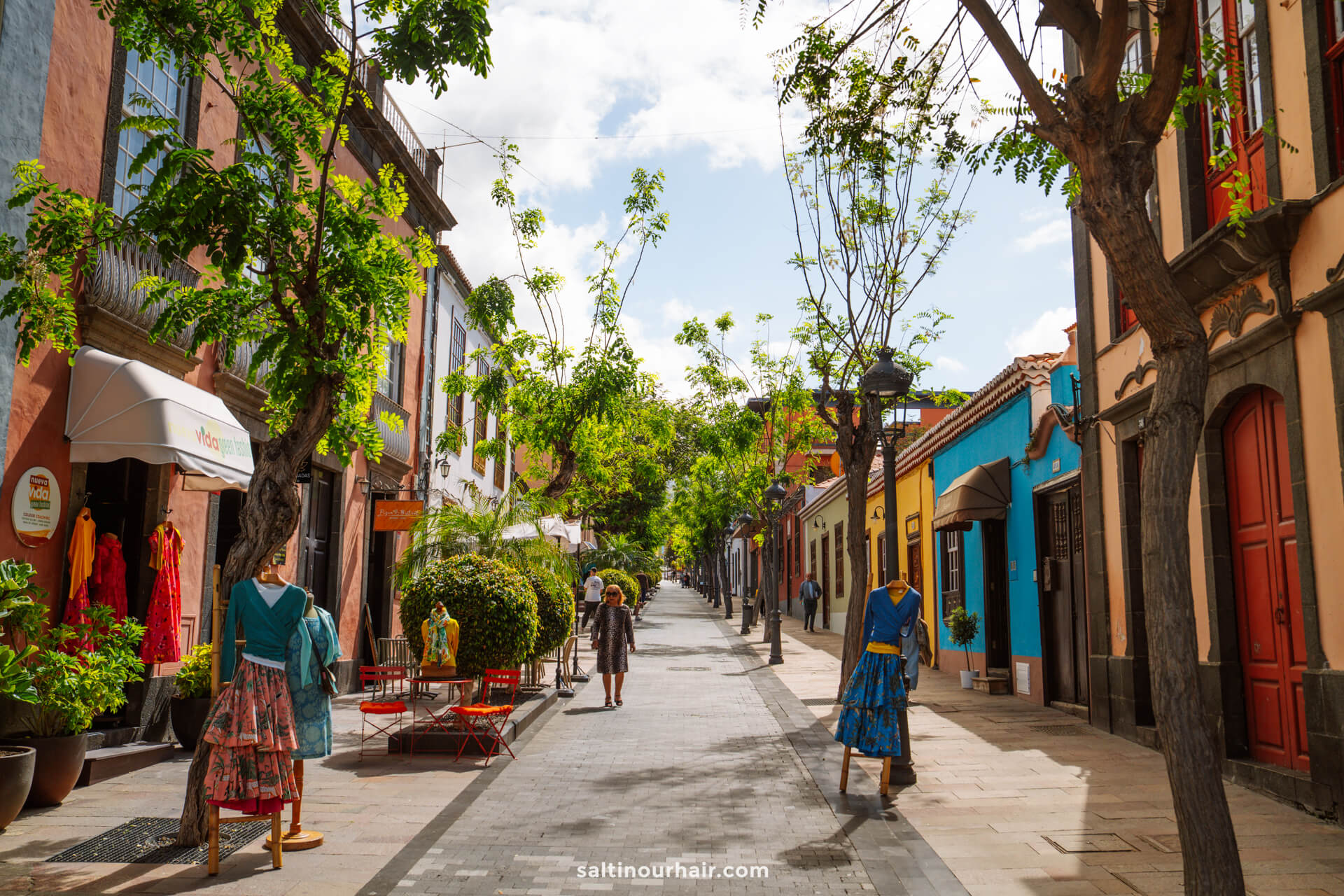 Pedestrian street in La Palma, Canary Islands, lined with colorful buildings, outdoor seating, mannequins in dresses, and trees; a few people stroll the pathway under a partly cloudy sky—a scene fit for any travel guide.